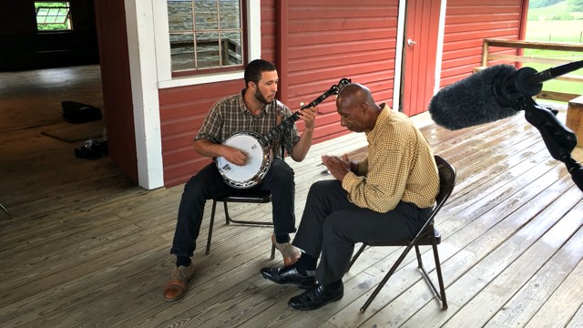 an older male clogging dancer seated on a rainy front porch with a banjo player, framed by sound equipment in the foreground