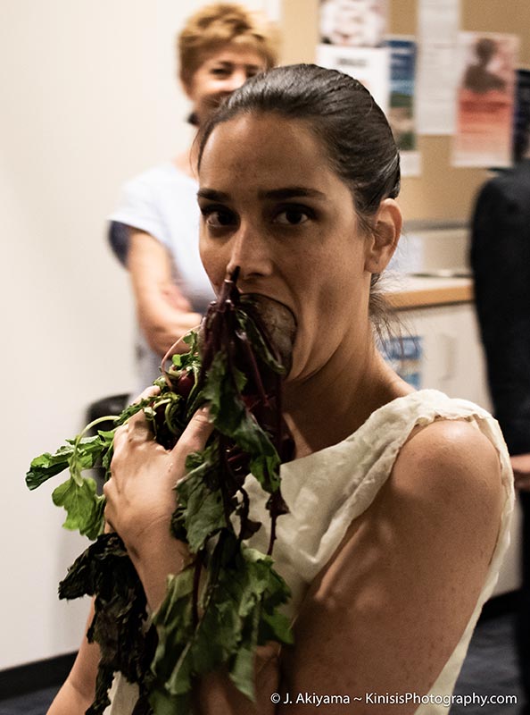 a woman stares down the camera defiantly, with her arms full of vegetables and a raw whole beet in her mouth