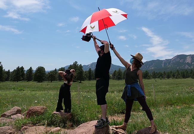 with the Boulder flatirons in the background, a dancer shifts her ribs one way and hips another with her hands behind her head, with one person filming, and another person holding an umbrella to shade the camera