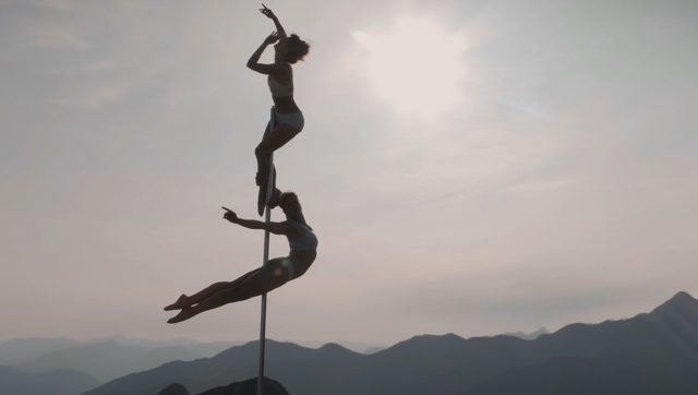 two dancers on a pole reach high and wide, set against a mountain backdrop