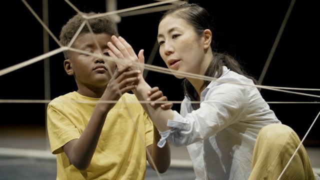 an Asian woman and a black boy dance with hands in a maze of string