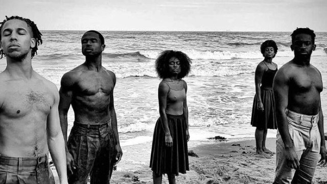 five dark skinned dancers stand in the sand at the beach with eyes closed