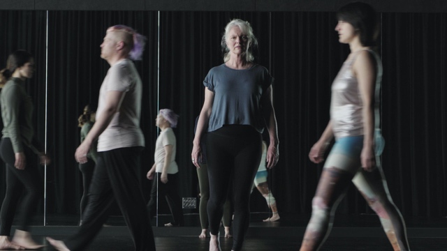 an older woman with white hair looks at the camera, while other dancers around her blur out of focus