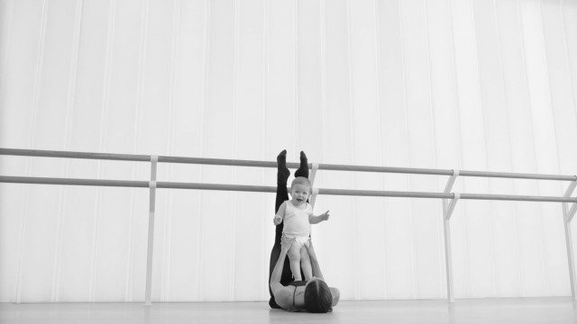 a ballerina holds a toddler on her chest while she stretches her legs on a bar in a dance studio