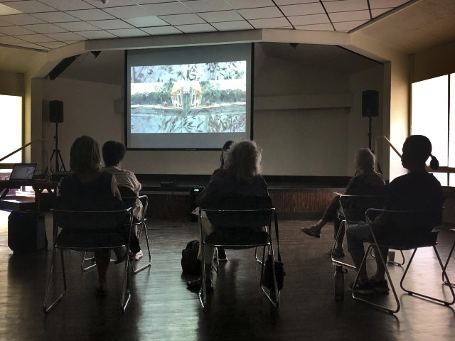several older adults sit in chairs, watching dance films projected onto a screen in the common area of their living center