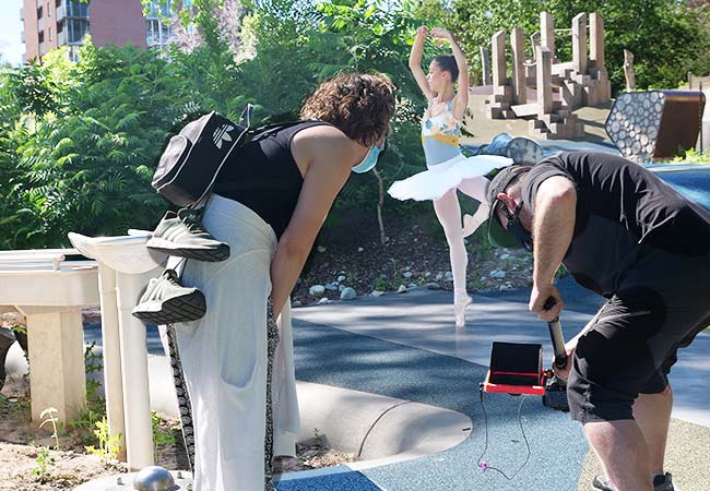 in the background a ballet dancer turns on pointe, while in the foreground a director and cinematographer eagerly watch the camera monitor