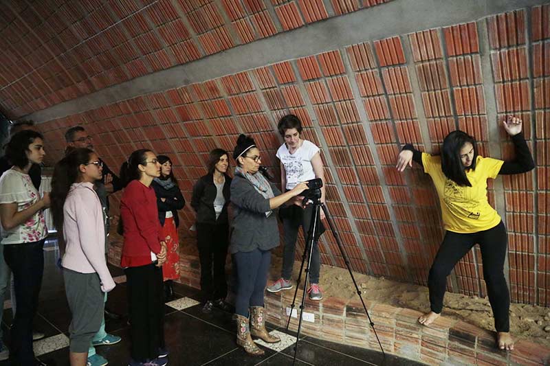 a dancer presses herself against a wall while a workshop facilitator and several participants watch from behind a camera on a tripod