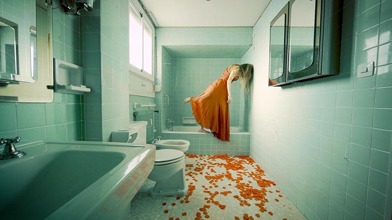 a woman dressed in orange, in a teal colored bathroom, with orange rose petals covering the floor