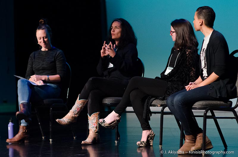 a dark haired woman describes an idea vividly with her eyes almost closed and hands in circular motion, surrounded by 3 other seated panelists on a stage