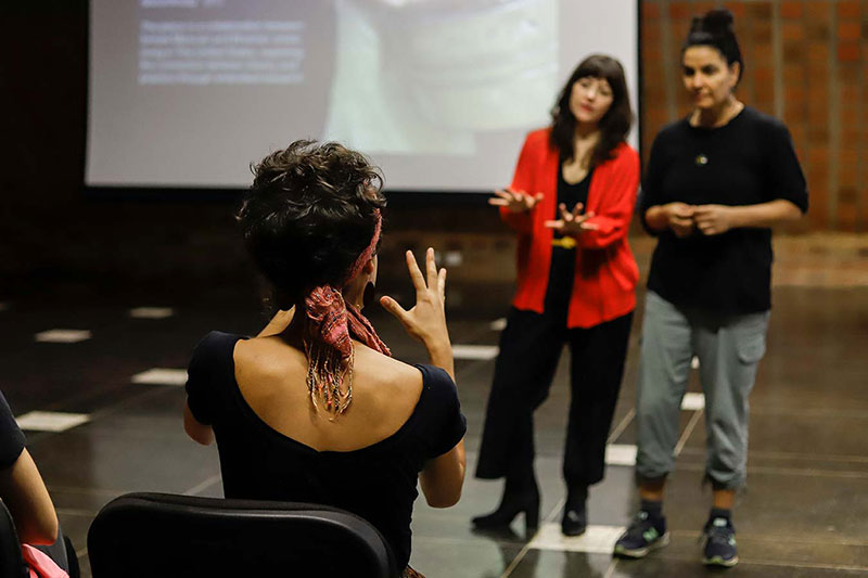 a seated workshop student explains vividly with her hands, while facilitator Ana Baer listens carefully to an interpreter