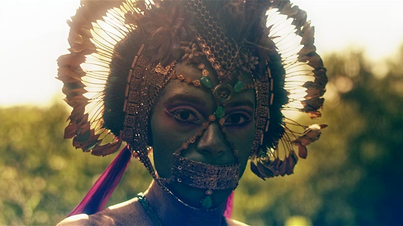 a dancer staring down the camera, though with eyes concealed by the gold coin-like features of a head dress that matches their gold plated feathers