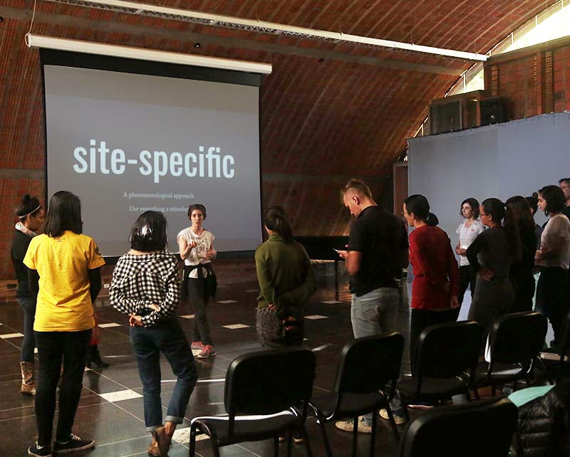 a woman leading a dance film workshop in Campinas, Brazil is facing the audience, standing in front of a projection screen that reads site specific in large letters