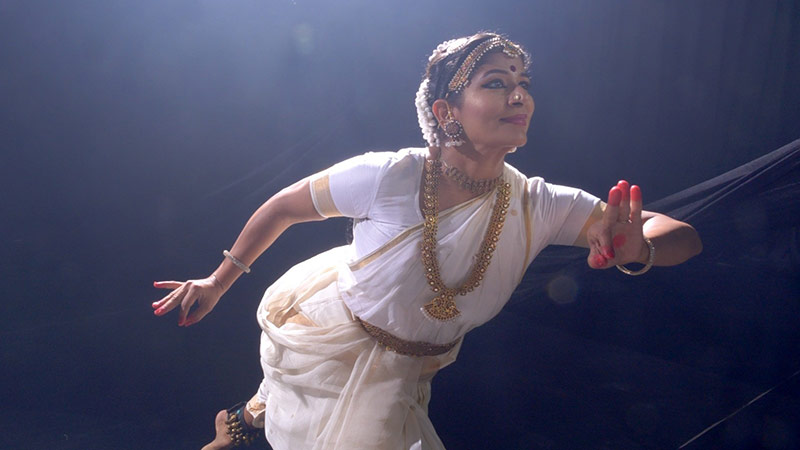 a woman in Hindi clothing, jewelry and makeup smiles, while extended in a deep lunge toward the camera