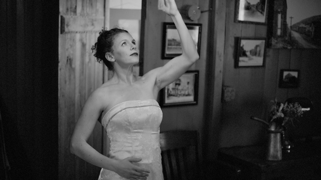 a woman in a wedding dress, seated in an old room, raises her left arm mechanically