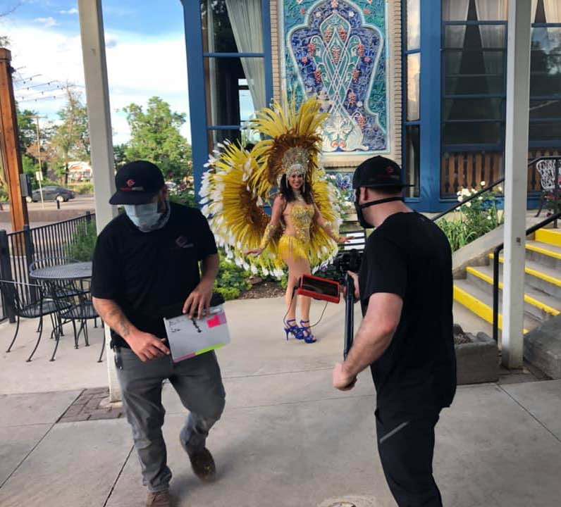 one man is walking away with the slate, while another is focusing his camera on a female samba dancer in fringe and feathers preparing to dance