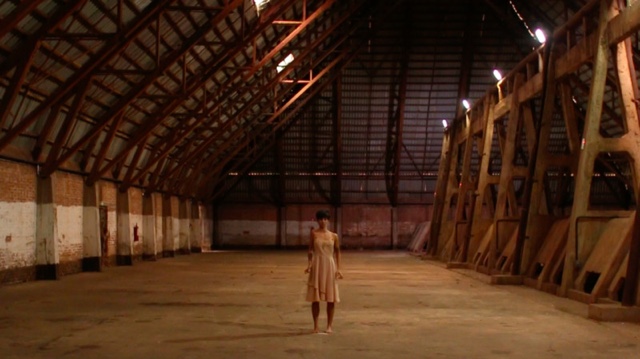 a woman with fair skin and short hair stands alone in a warehouse