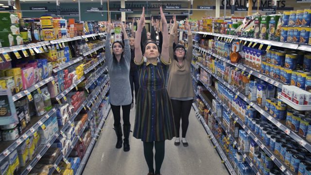 several dancers reach their arms upward, filling a grocery store aisle