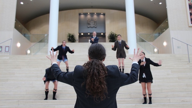 several women in men's suit jackets, ties, and underwear, on the steps of a building gesture wildly