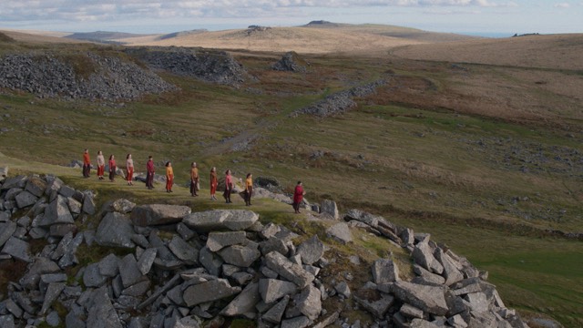a line of people stand on a rocky cliff, surrounded by hills