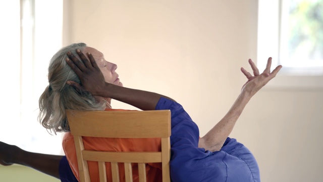 an older white woman in a chair closing her eyes, with a younger black woman touching her face