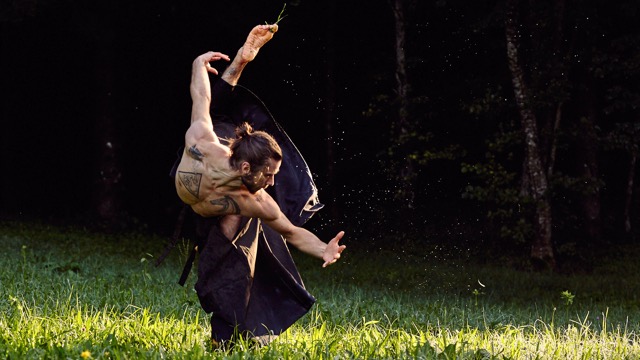 a bare chested white male dancer reaches toward the grass with a leg thrown up behind him