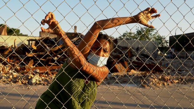 a brown-skinned woman with a mask hangs on and looks through a fence