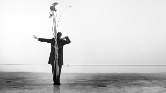 a white man in a tux with a sculpture of dead flowers tied to his back