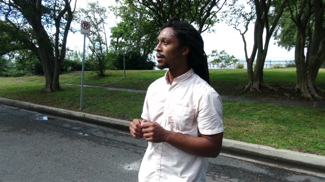 a man with dark skin and dreadlocks in a collared shirt standing on a neighborhood street