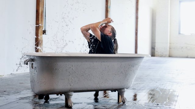 a light skinned dancer in a t-shirt in a bathtub flings water onto her face