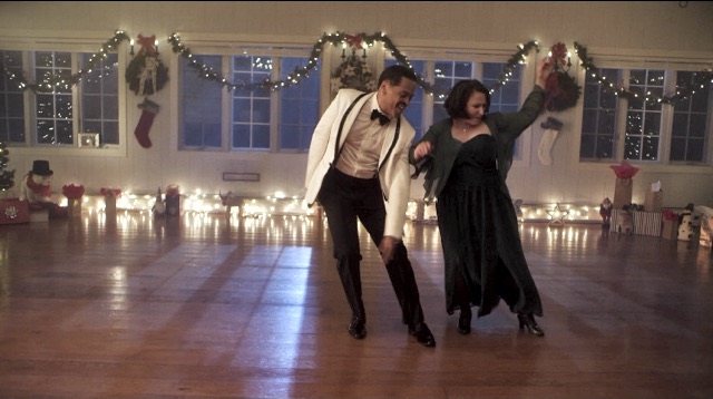 a man and woman tap dance in a large room with bare floor and Christmas decorations