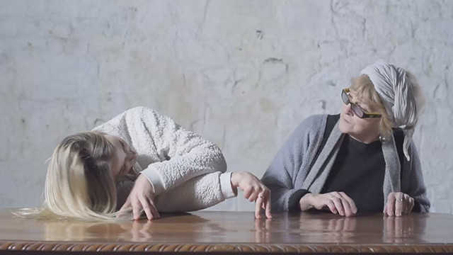 two women sit at a table; one uses her fingers to mimic a dancer's legs