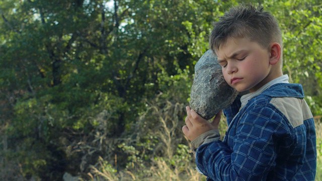 a young boy holds a rock to his ear with eyes closed