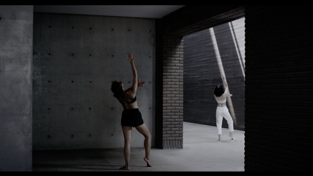 two women reach one arm forward and one arm up, separated by a brick archway and strikingly different lighting