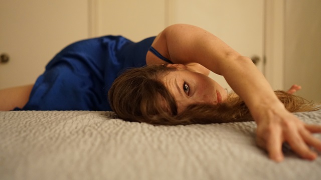 a woman in a blue dress lays on her side, reaching and looking toward the camera