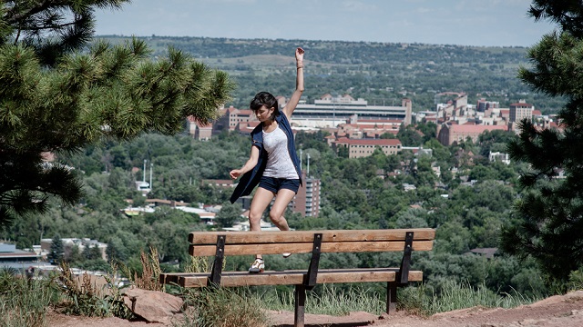 a light skinned dancer with short black hair swings her arms while tapping on a bench overlooking a city
