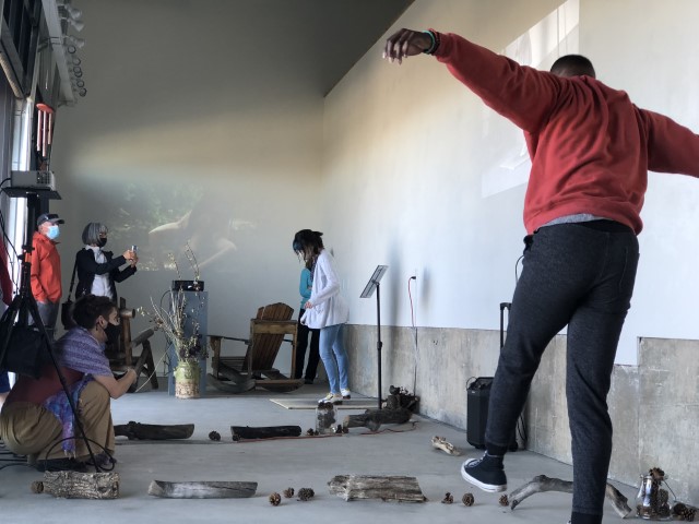 a dark skinned man in an orange sweatshirt dances in the foreground while a woman with black hair taps on a tap board in the background