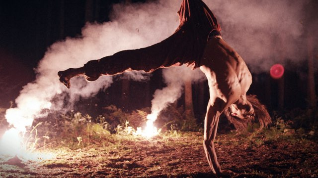 a shirtless man performs a one-handed cartwheel in a forest illuminated by road flares