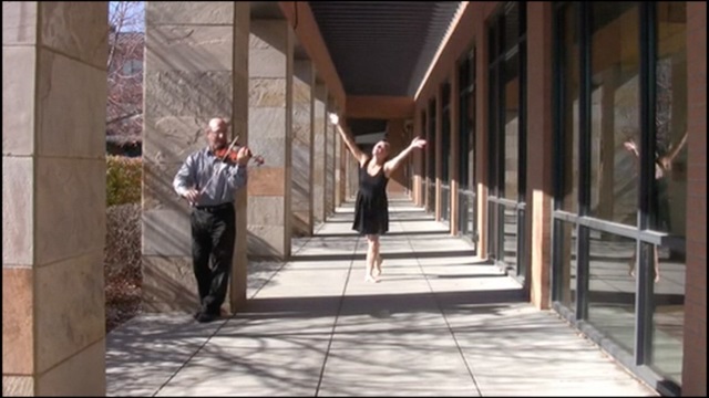 a woman in a black dress dances joyfully outside a building while a man plays a violin
