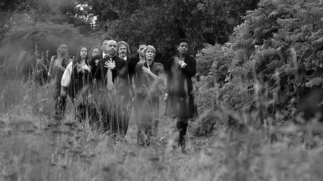 a group of dancers walk through the woods with their left hands placed over their chests