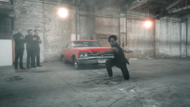 a man dressed in black dances on one knee in front of a bright red 1950s car while three other men in black watch from the background