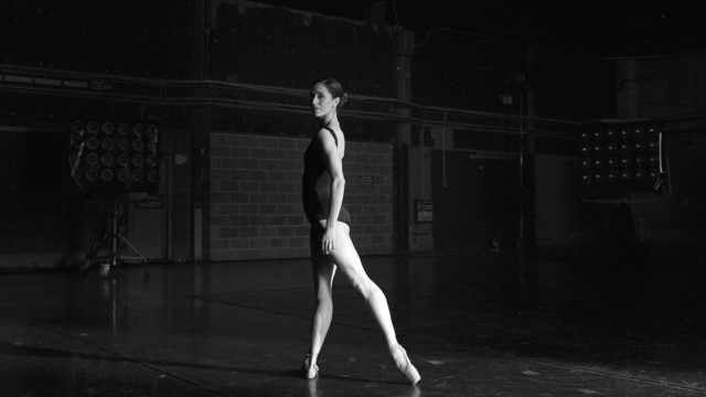 a light skinned ballerina in a dark room looks over her left shoulder and toward us