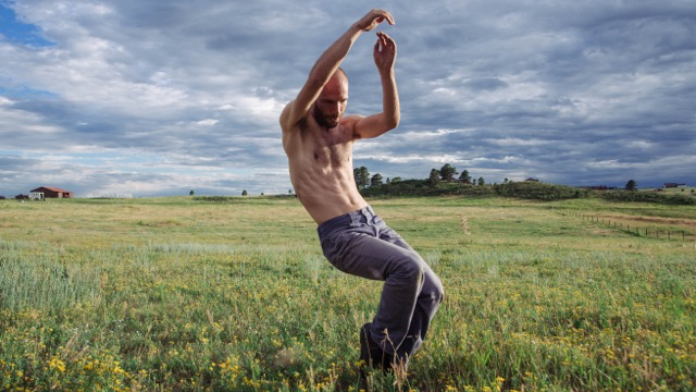 a shirtless male dancer in a field balanced on toes with bent knees