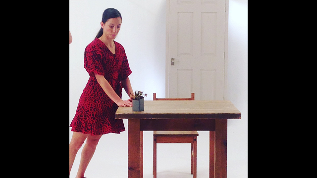 a woman with dark hair and a red dress leans gently forward onto a square table with a small potted plant