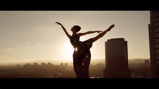 a female dancer silhouetted by the setting sun poses atop a building with a city in the background