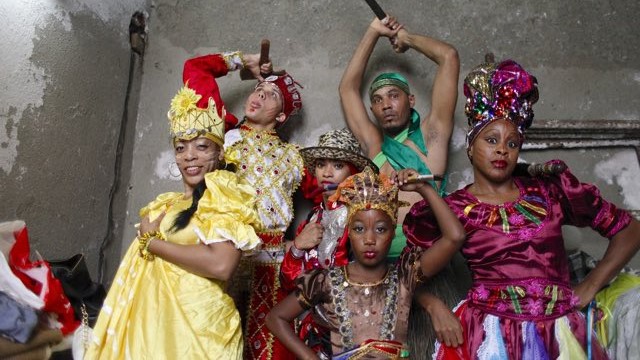 six dancers with elaborate, colorful costumes and a range of skin tones pose for the camera