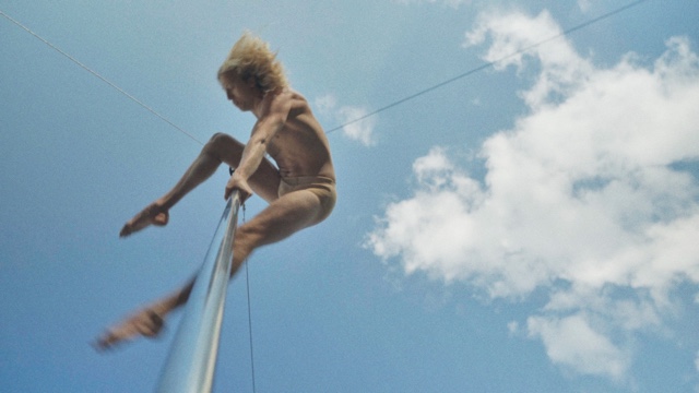 a shirtless man performs on a tall metal pole as seen from a camera positioned at its base