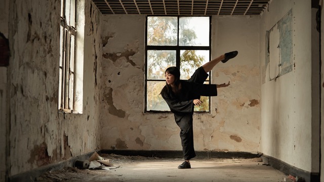 a woman dressed in loose black clothing dances in a old room with two windows and peeling paint