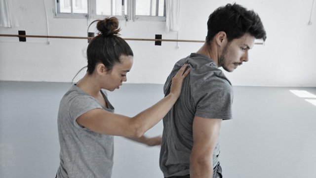 in a dance studio, a woman places her hand on the upper back of a man with a mustache