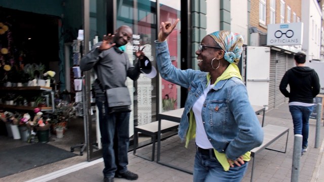 a joyful looking woman in a jean jacket raises her finger in the air while walking past an open shop on a city street