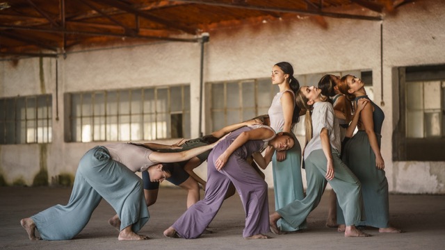 seven dancers in loose, flowy clothing, perform in a warehouse